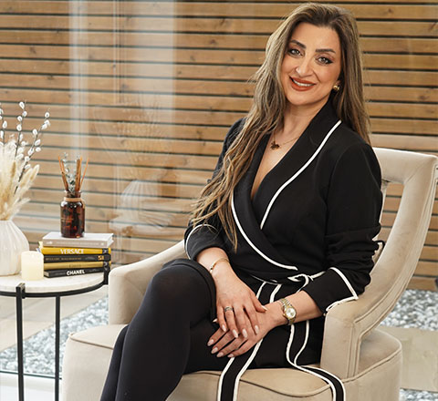 woman sitting elegantly in a modern chair smiling wearing a stylish black outfit with white accents two books on a side table decorative plant in background